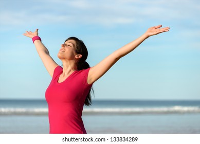 Blissful fitness woman raising arms to the sky on beach at sunset after working out. Happy female athlete feeling the breeze on sea background. Achievements and goals on sport concept.