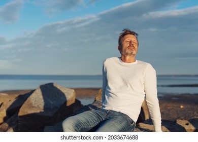 Blissful Man Taking A Moment To Relax And Meditate At Sunset Raising His Face To The Sun As He Relaxes On Rocks On A Tropical Beach With Ocean Backdrop