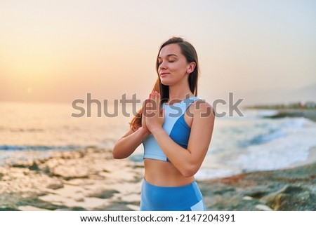 Сoncentrated blissful one fitness woman with closed eyes and praying hands standing alone with meditation position while breathing exercises by sea. Mental mind care and healthy habits