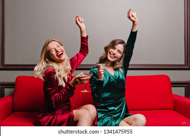 Blithesome girls in velvet dressed drinking wine. Indoor portrait of glamorous ladies sitting on sofa with glasses of champagne.