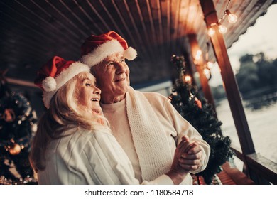 Blithesome old couple wearing red caps is dancing on porch and looking aside with dreamy smile. Decorated trees at background. Copy space on right side - Powered by Shutterstock