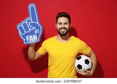 Blithesome young bearded man football fan in yellow t-shirt cheer up support favorite team look camera hold soccer ball fan foam glove finger up isolated on plain dark red background studio portrait