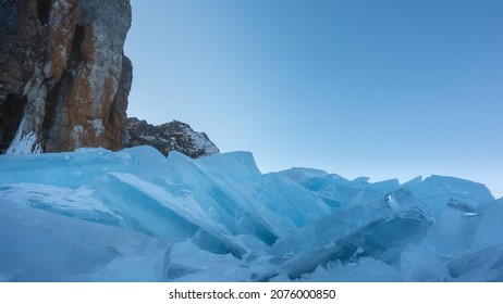 A block of blue hummocks against an azure sky. Thick ice floes lie in disarray at the base of a granite rock. Lake Baikal