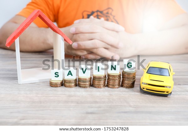 Block letters on top of\
stack of coins and a house model and a car model with hand of a man\
at the back 