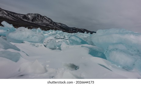 A block of turquoise ice hummocks lies in disarray on the snow. A mountain range against a cloudy sky. Lake Baikal in winter