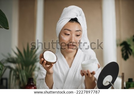 Blogger or content creator asian woman in towel and bathrobe preparing natural cosmetics at home holding coconut homemade cream in glassware for skin and hair care home spa