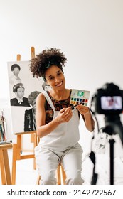 Blogger live streaming from her studio. Happy young female painter smiling while recording a painting tutorial with a camera. Young female freelancer creating a video blog in her art studio.