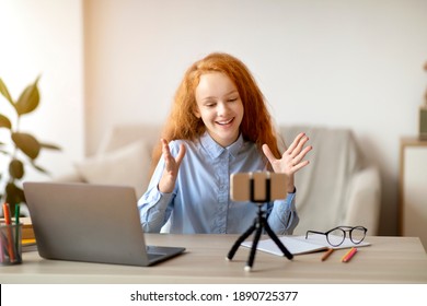 Blogging Concept. Smiling red-haired teen girl recording lifestyle blog talking to camera of mobile phone on tripod, sitting at desk at home. Happy young influencer filming vlog for her channel