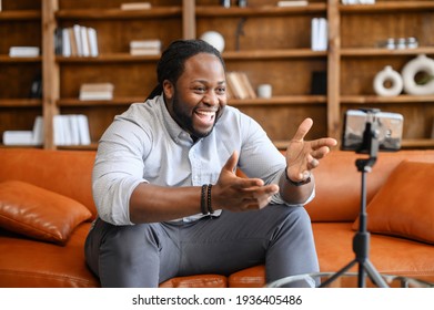 Blogging concept. Young African American male blogger in casual clothes streaming video online or recording tutorial for social media network, looking at the mobile phone on a tripod and laughing - Powered by Shutterstock