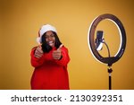 blogging, new year, technology and people concept - smiling african-american girl vlogger in a red hoodie and a Santa hat with a lighted ring lamp and phone showing thumbs up on a yellow background