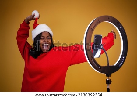 blogging, new year, technology and people concept - laughing african-american girl vlogger in a red hoodie and a Santa hat with a lighted ring lamp and phone holding a pompom on a yellow background