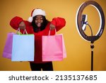 blogging, shopping, new year, technology and people concept - Smiling young African-American girl in a Santa hat with a ring lamp showing colorful packages on a yellow background with copy space