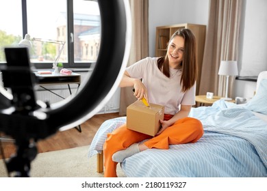 blogging, technology and people concept - happy smiling girl blogger with ring light and smartphone unpacking parcel box with knife at home