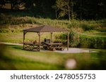 Blome, Latvia - September 11, 2023 - A wooden gazebo with a picnic table and barbecue grill by a pond, surrounded by greenery and tall grass, offering a serene outdoor setting.