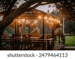 Blome, Latvia - September 11, 2023 - A cozy outdoor gazebo with hanging plants and string lights at dusk, surrounded by greenery, creating a warm and inviting atmosphere