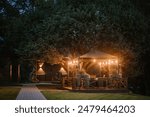 Blome, Latvia - September 11, 2023 - A cozy gazebo illuminated by string lights at dusk, surrounded by lush trees and a well-kept lawn, creating a warm and inviting atmosphere.