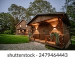 Blome, Latvia - September 11, 2023 - Two wooden cabins with illuminated exteriors, surrounded by greenery and paved walkways, nestled under large trees in a serene setting.