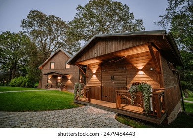 Blome, Latvia - September 11, 2023 - Two wooden cabins with illuminated exteriors, surrounded by greenery and paved walkways, nestled under large trees in a serene setting. - Powered by Shutterstock
