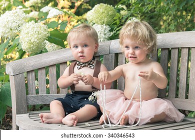 Blond Baby Boy And Girl In Formal Dress Sitting On Wooden Bench In A Beautiful Garden With White Flowers, She Wears Light Pink Skirt And He Bow-tie