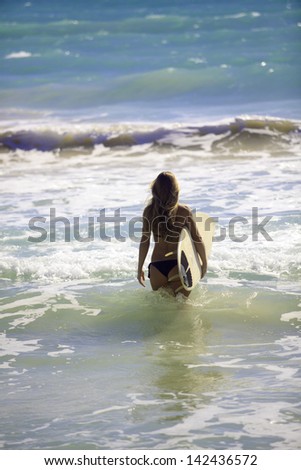 blond in bikini with her surfboard heading into the waves in hawaii