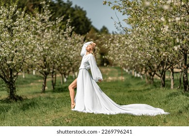 Blond blooming garden. A woman in a white dress walks through a blossoming cherry orchard. Long dress flies to the sides, - Powered by Shutterstock