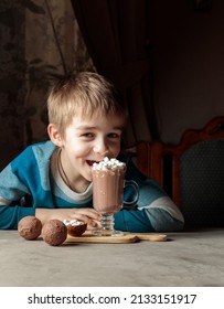 A Blond Boy Eats Marshmallows From A Glass Of Cocoa Or Hot Chocolate. Portrait, Low Key.