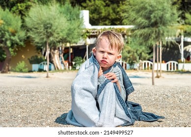 Blond boy frowns wrapped with towel sitting on beach after swimming. Toddler rests at bright sunlight during holidays in Omish of Croatia closeup