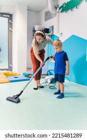 Blond Boy And His Teacher Cleaning The Floor With The Vacuum Cleaner In The Nursery, Vertical Shot. High Quality Photo