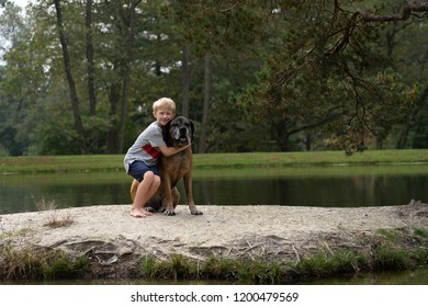 Blond Boy Is Hugging A Large Dog, English Mastiff, Standing On A Small Island Of A Lake Or A  Pond. Boy Is Smiling, Sunny Warm Day. Wide Shot. 