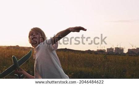 A blond boy launches a toy glider plane into the sky on a field, the setting sun beautifully fills the space with evening light. Buildings are being constructed in the distance.