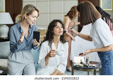 Blond Bridesmaid With Smartphone Showing Online Photos Of Wedding Makeup To Brunette Girl And Visage Artist Applying Powder On Her Face