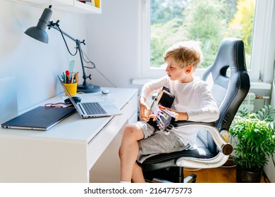 Blond Caucasian Boy Having Music Online Lesson Play On Guitar Using Laptop Sitting On Desk At Home. Online Learning