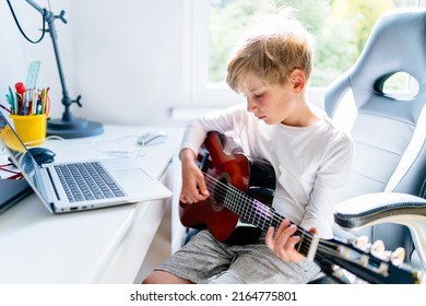 Blond Caucasian Boy Having Music Online Lesson Play On Guitar Using Laptop Sitting On Desk At Home. Additional Online Education