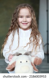 Blond Caucasian Female Child With Dark Narrow Eyes Indoors Holding Soft Toy And Looking Cheerfully Smiling. Wearing White T-shirt. Studio Portrait. Happy Childhood Concept. Candid Authentic Image