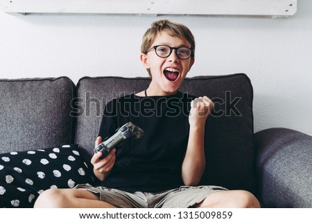Blond child with eyeglasses legs crossed playing video games sitting on sofa  at home legs crossed Young student resting play on line with friends Domestic pastime challenge web exult happy winning