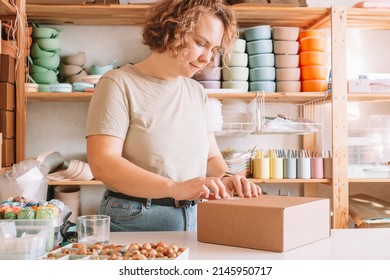 Blond confident woman packing and wrapping parcel with silicone baby dishes in cardboard box package, stamping cardboard. Colorful skein of thread, beads on desk. Workshop store, small business