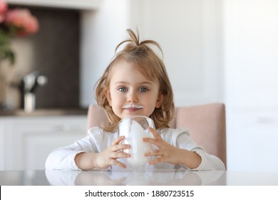 Blond Cute Baby Girl With Blue Eyes With Traces Of Milk On The Lips Is Holding A Glass Of Milk Siting At A White Table In A White Kitchen. Milk For Good Health