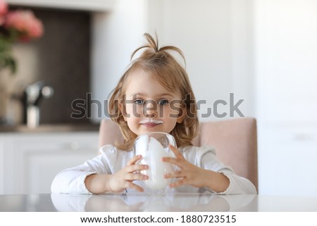 Blond cute baby girl with blue eyes with traces of milk on the lips is holding a glass of milk siting at a white table in a white kitchen. Milk for good health