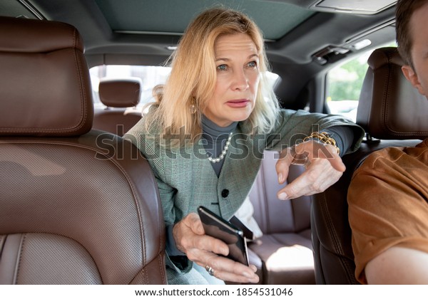 Blond elegant mature businesswoman showing taxi\
driver where to go and asking him something while sitting on\
backseat of car in the\
morning