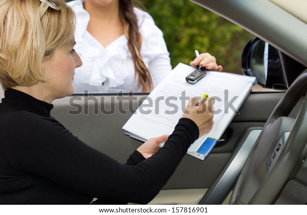 Blond female driver signing the deal on the\
purchase of a new car on the contact being held through the open\
window by the saleslady