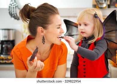 Blond Girl In Bat Costume With Happy Mother In Decorated Kitchen Eating Freshly Homemade Halloween Biscuits For Trick Or Treat. Traditional Autumn Holiday