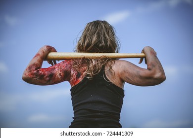 Blond girl in a black tank top and a burnt, bloody shoulder (practical special effect), looking up at the blue sky with a baseball bat in her hands