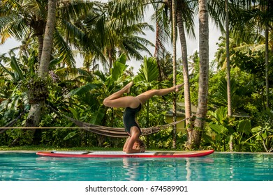 Blond Girl Doing A SUP Yoga Headstand In A Pool In A Ceylon Jungle.
