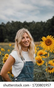 A blond girl on a sunflower field with a denim overall and white tee smiling
