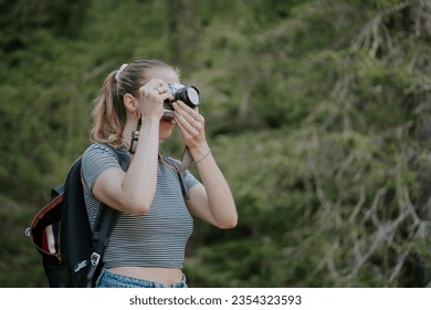 Blond girl photographs with an analog camera. Young hiker passionate about photography frames the scene. Woman with hair in a ponytail, striped t-shirt and backpack.