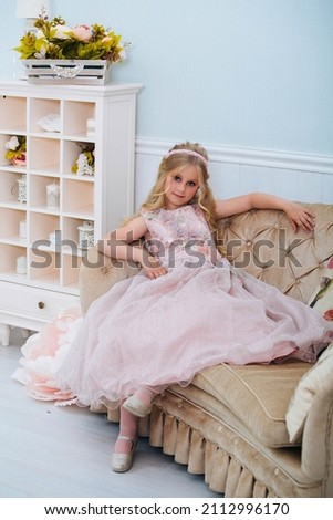 Blond hair beautiful little girl in designer black dress outfit sits inside in room with flower and stairs background holding lamp