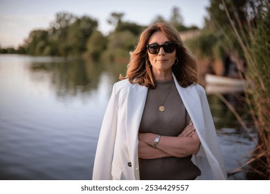 A blond haired middle-aged woman standing on the pier by the lakeside. Attractive female wearing white suit and sunglasses.  - Powered by Shutterstock