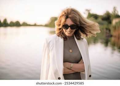 A blond haired middle-aged woman standing on the pier by the lakeside. Attractive female wearing white suit and sunglasses.  - Powered by Shutterstock