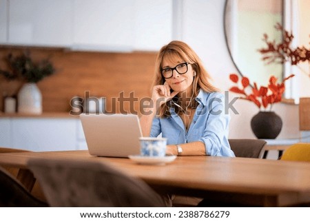Blond haired woman sitting at home and usning laptop and earphone for work. Home office. Confident businesswoman having web conference or browsing on the internet. 