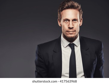 Blond Handsome Young Man Looking At Camera With A Serious And Determined Facial Expression While Wearing Business Black Suit, White Shirt And Tie, Portrait With Copy Space On Grey